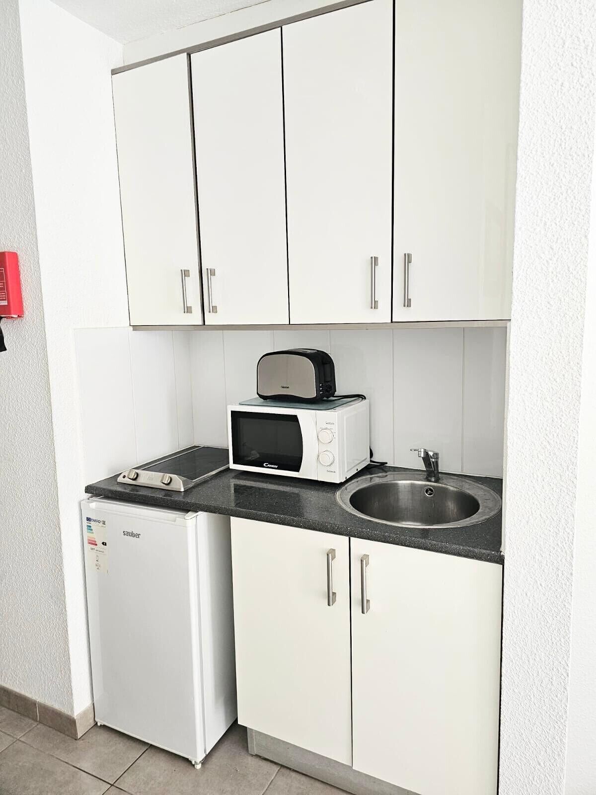 Een kleine keuken met witte kastjes, een magnetron, een broodrooster en een spoelbak.