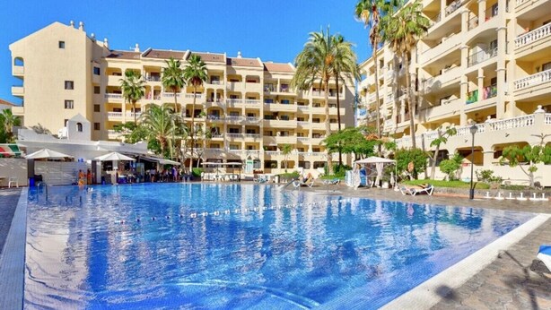 A large swimming pool with clear blue water, surrounded by palm trees and a multi-story hotel building with balconies. - The Poolside Haven (Los Cristianos)