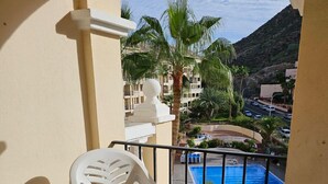 A balcony with a view of a swimming pool, palm trees, and a mountainous landscape. - The Poolside Haven (Los Cristianos)