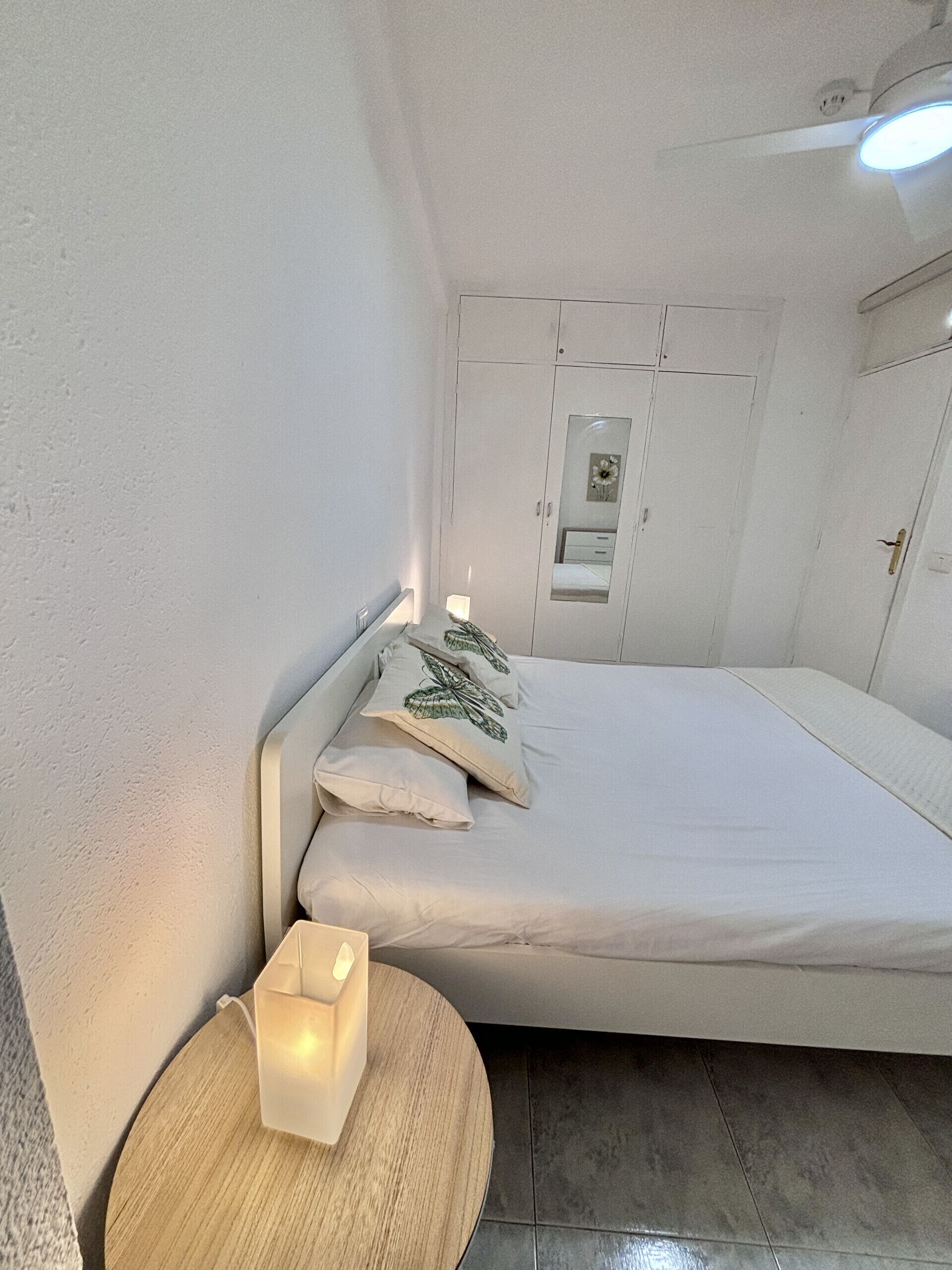 Een kleine, verzorgde slaapkamer met een bed, een nachtkastje met een kaars en een garderobe met spiegel.