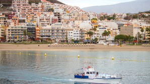 A coastal town with a boat on the water, buildings in the background, and a sandy beach. - Sunny Views Los Cristianos (Los Cristianos)