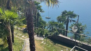 A balcony with a view of a calm lake, surrounded by palm trees and a stone pathway. - Villa Angelina (Cannero Riviera)
