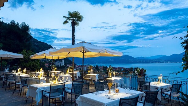 An outdoor dining area with tables and chairs, umbrellas, and a view of a lake and mountains. - Villa Angelina (Cannero Riviera)
