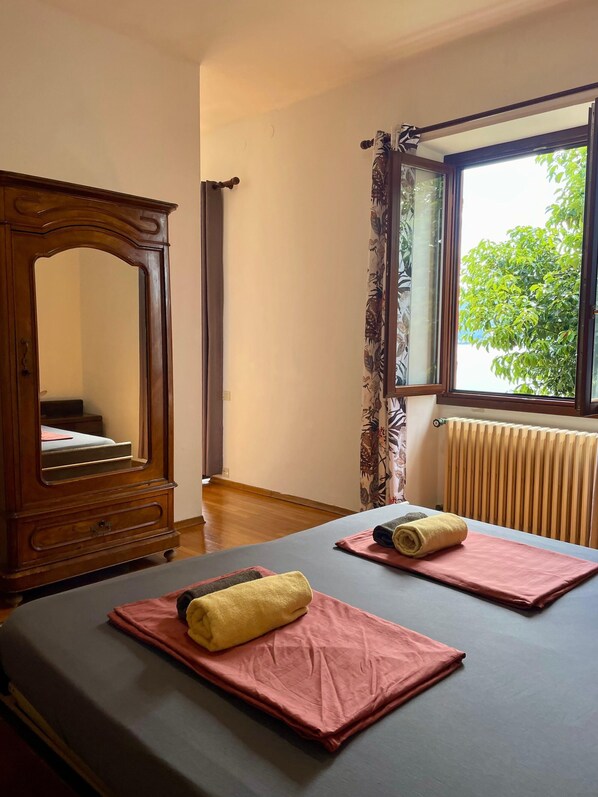 A hotel room with two beds, a large mirror, a window with curtains, and a radiator. - Villa Angelina (Cannero Riviera)