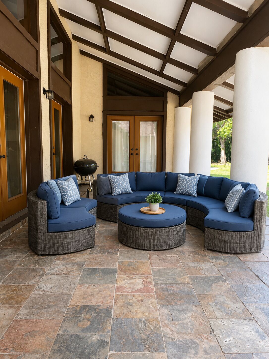 A covered outdoor seating area with a curved sectional sofa and round ottoman, stone flooring, and a view of a building with large windows and a door.