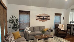 Ein Wohnzimmer mit grauem Sofa, einem Couchtisch und einer Pflanze. Ein Fenster mit Jalousien und ein Gemälde an der Wand sind zu sehen.