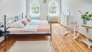 Un dormitorio con una cama, un tocador, un sillón mecedora y dos ventanas.