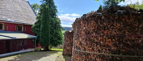 A red building with a tiled roof, a large tree in front, and stacks of firewood.