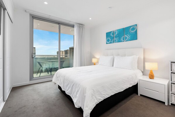 A hotel room with a large bed, two bedside tables with lamps, a view of the city through a sliding glass door, and a blue abstract painting on the wall. - Adelaide Central Skyline Panoramic Suite Two (Adelaide)