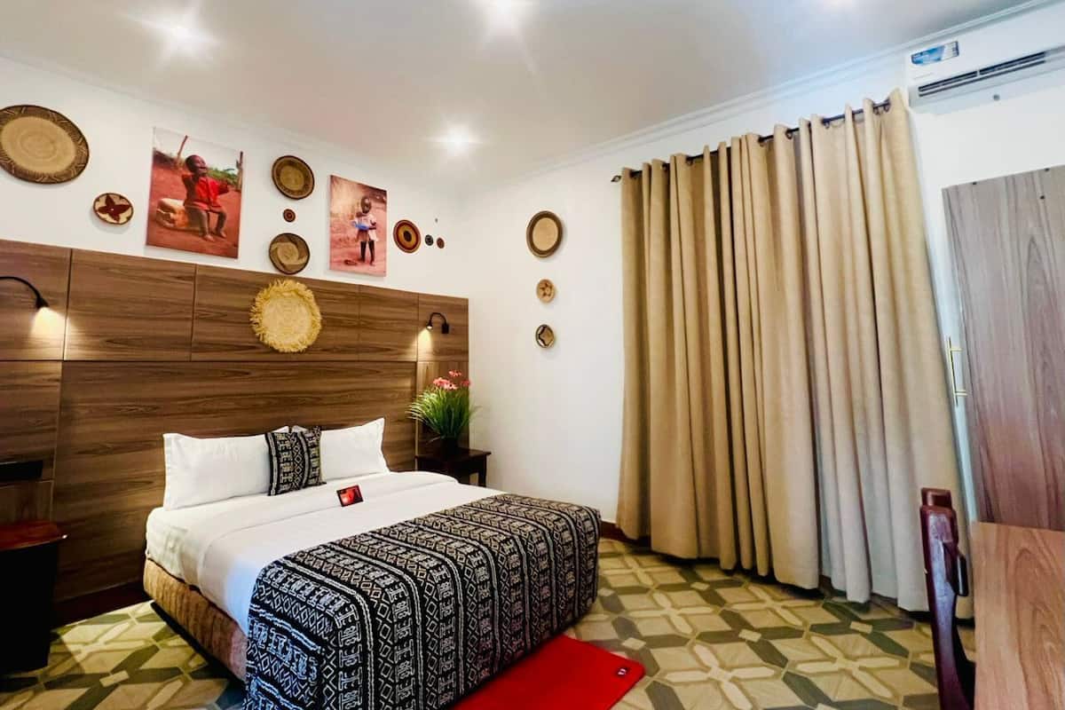 Habitación de hotel con cabecera de madera, cama con ropa de cama estampada, alfombra roja y un ventanal con cortinas.
