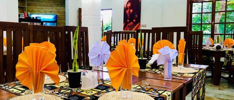 A restaurant with wooden chairs and tables, decorated with orange napkins and a potted plant.
