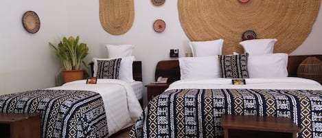 A hotel room with two beds, each with patterned bedding, a large woven wall hanging, a potted plant, and a wooden nightstand with a phone.