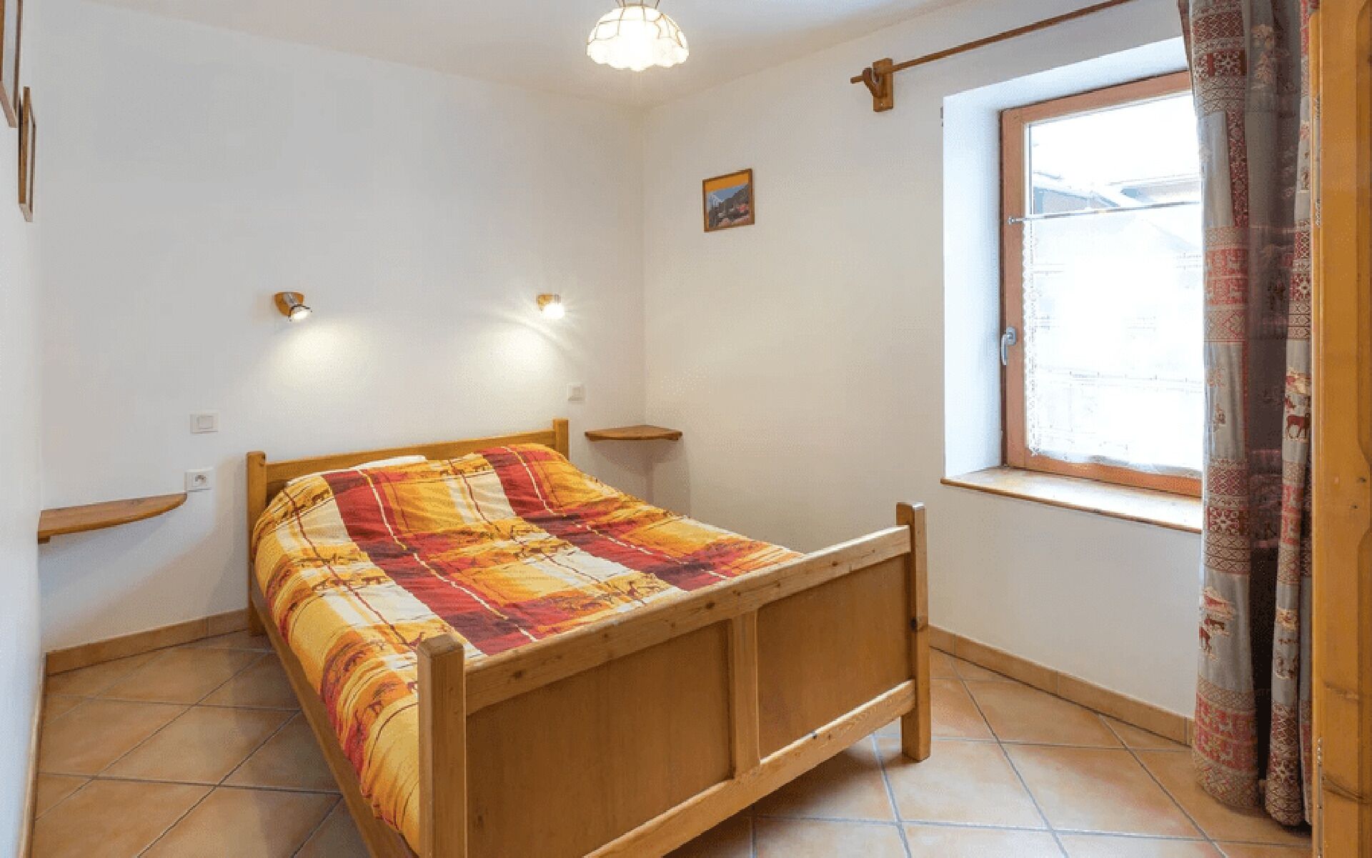 A single-bed room with a wooden frame, a colorful bedspread, two wall-mounted lights, a small shelf, a window with curtains, and a picture on the wall.