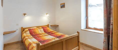 A single-bed room with a wooden frame, a colorful bedspread, two wall-mounted lights, a small shelf, a window with curtains, and a picture on the wall.