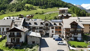A cluster of traditional chalet-style buildings with stone facades and wooden balconies, set against a backdrop of lush green hills and a clear blue sky. - Gites Simond welcomes you to Arvieux in the heart of the Queyras Regional Park, (ARVIEUX)