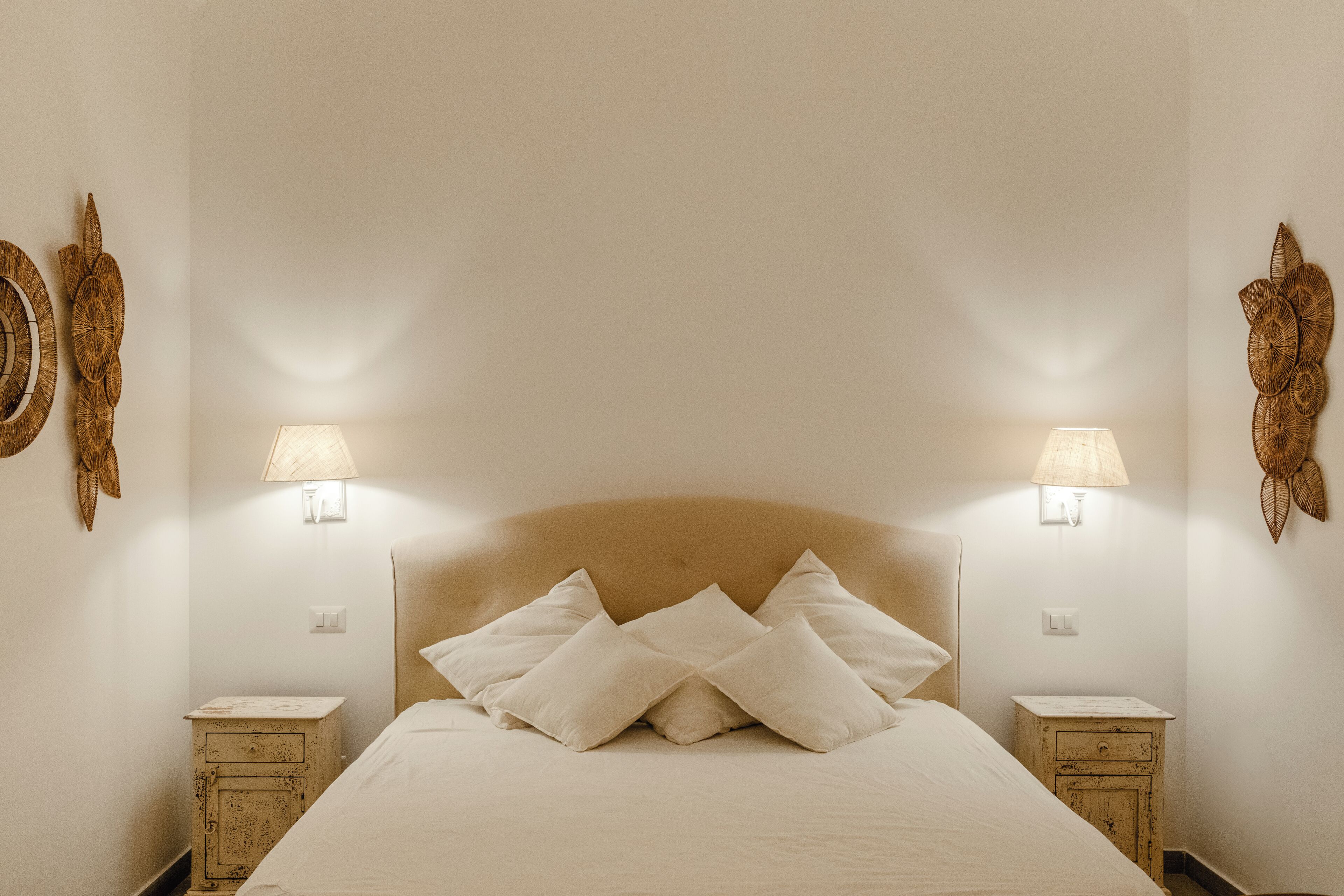 Un lit bien fait, avec du linge de lit blanc et des oreillers, flanqué de deux tables de chevet surmontées de lampes, dans une pièce aux murs sobres et agrémentée d’œuvres d’art décoratives.