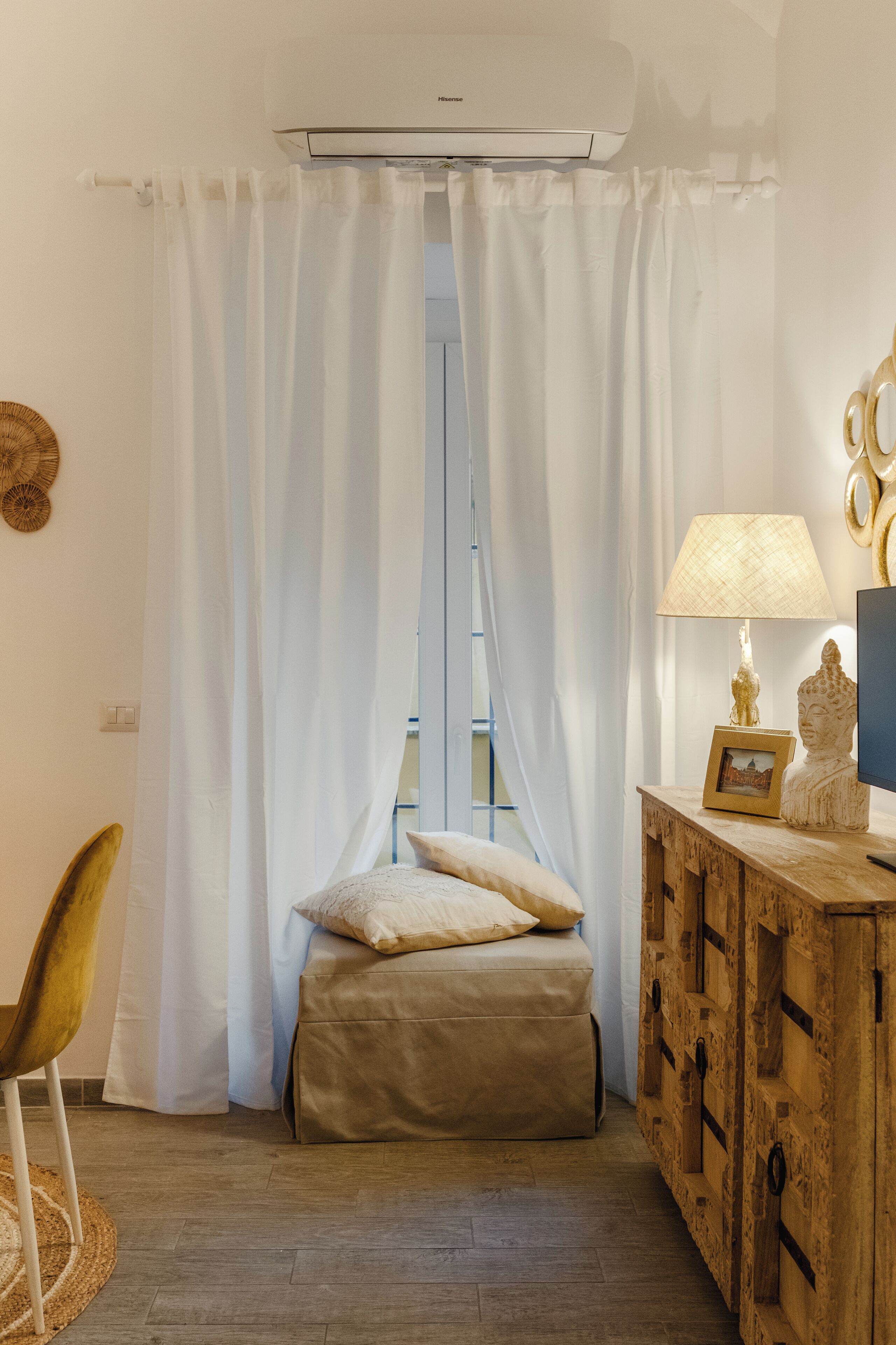 Quarto com cama, cómoda de madeira, luminária e janela com cortinas brancas.