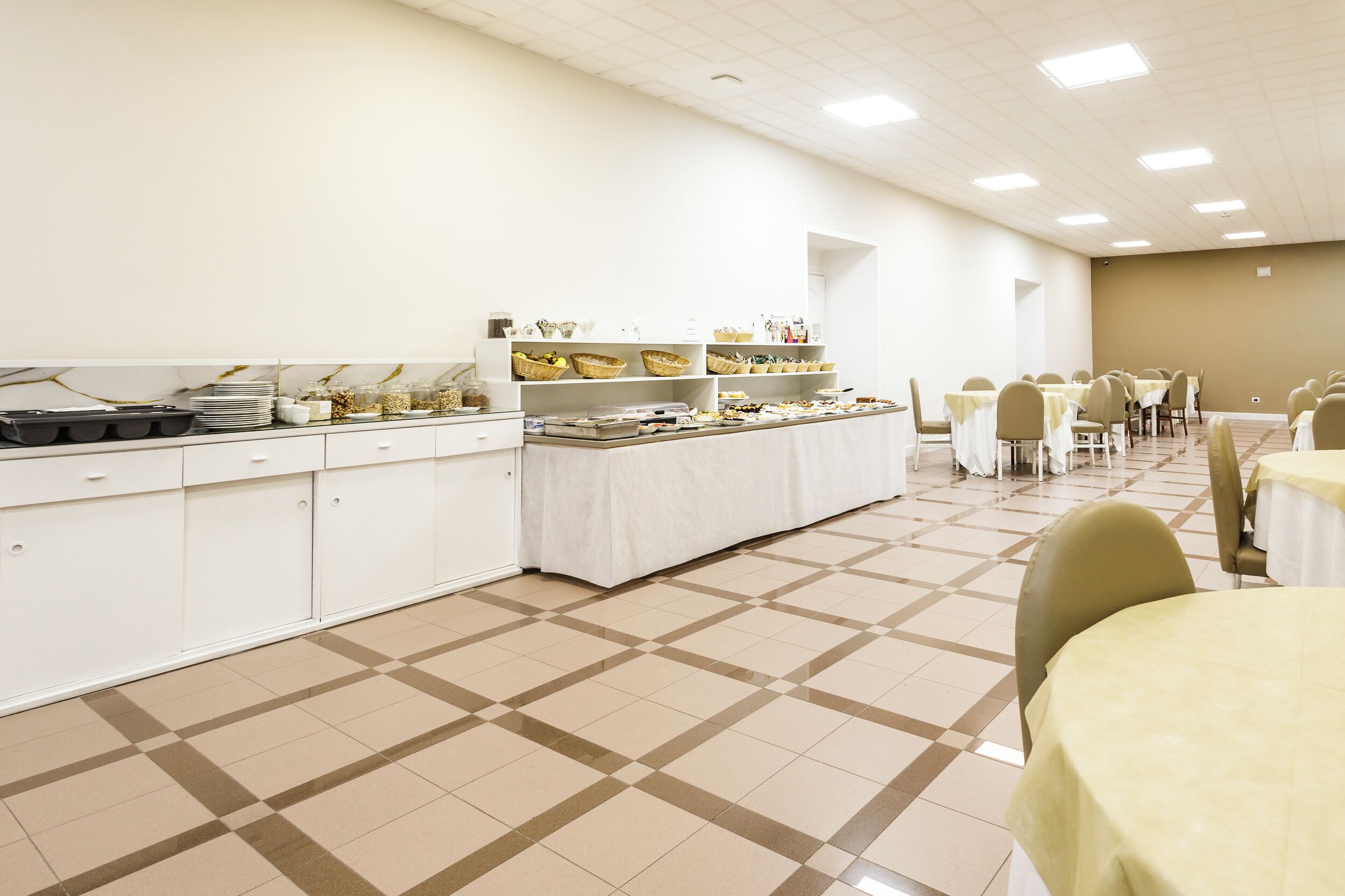 Un buffet avec des tables et des chaises blanches, un long comptoir où sont présentés les plats, et un sol carrelé.