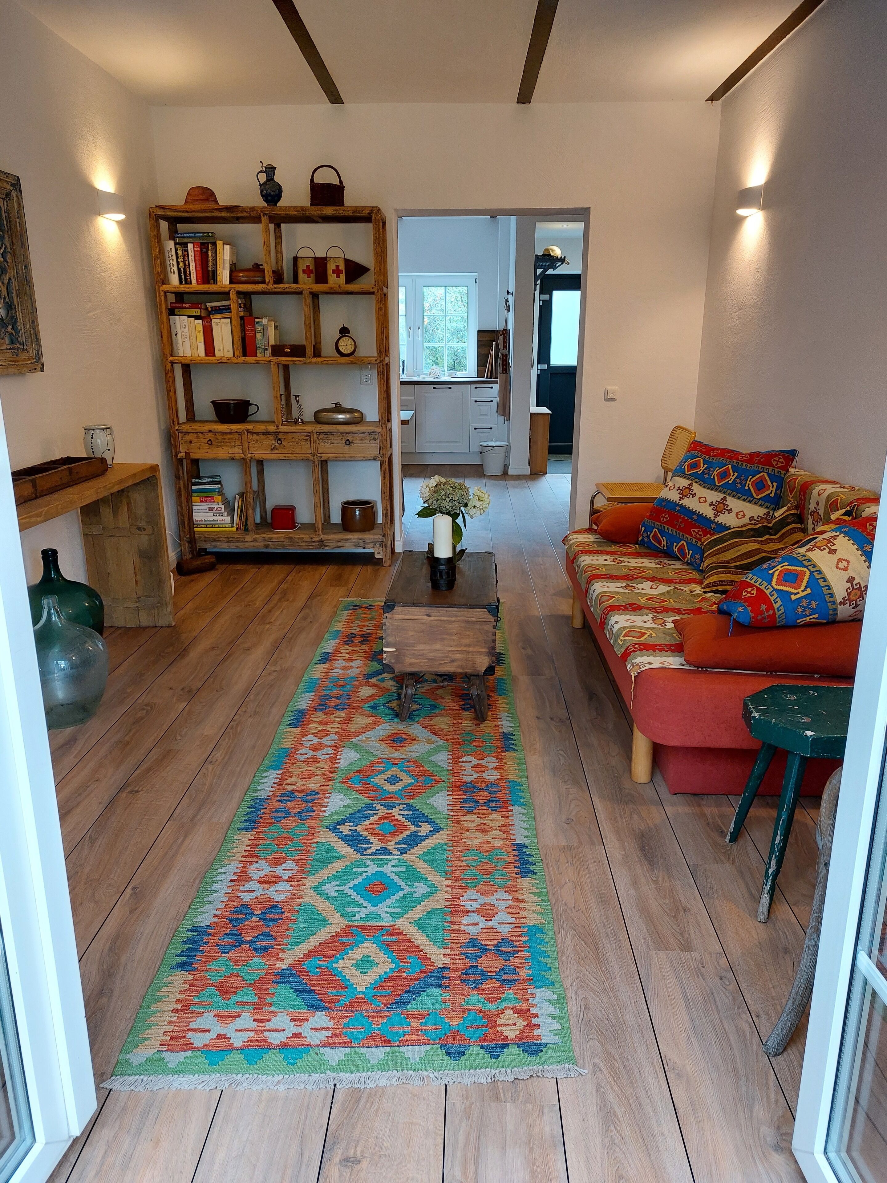 Ein gemütliches Wohnzimmer mit Holzboden, einem farbenfrohen Teppich, einer Couch mit gemusterten Kissen und einem Bücherregal voller Bücher und Dekoration.