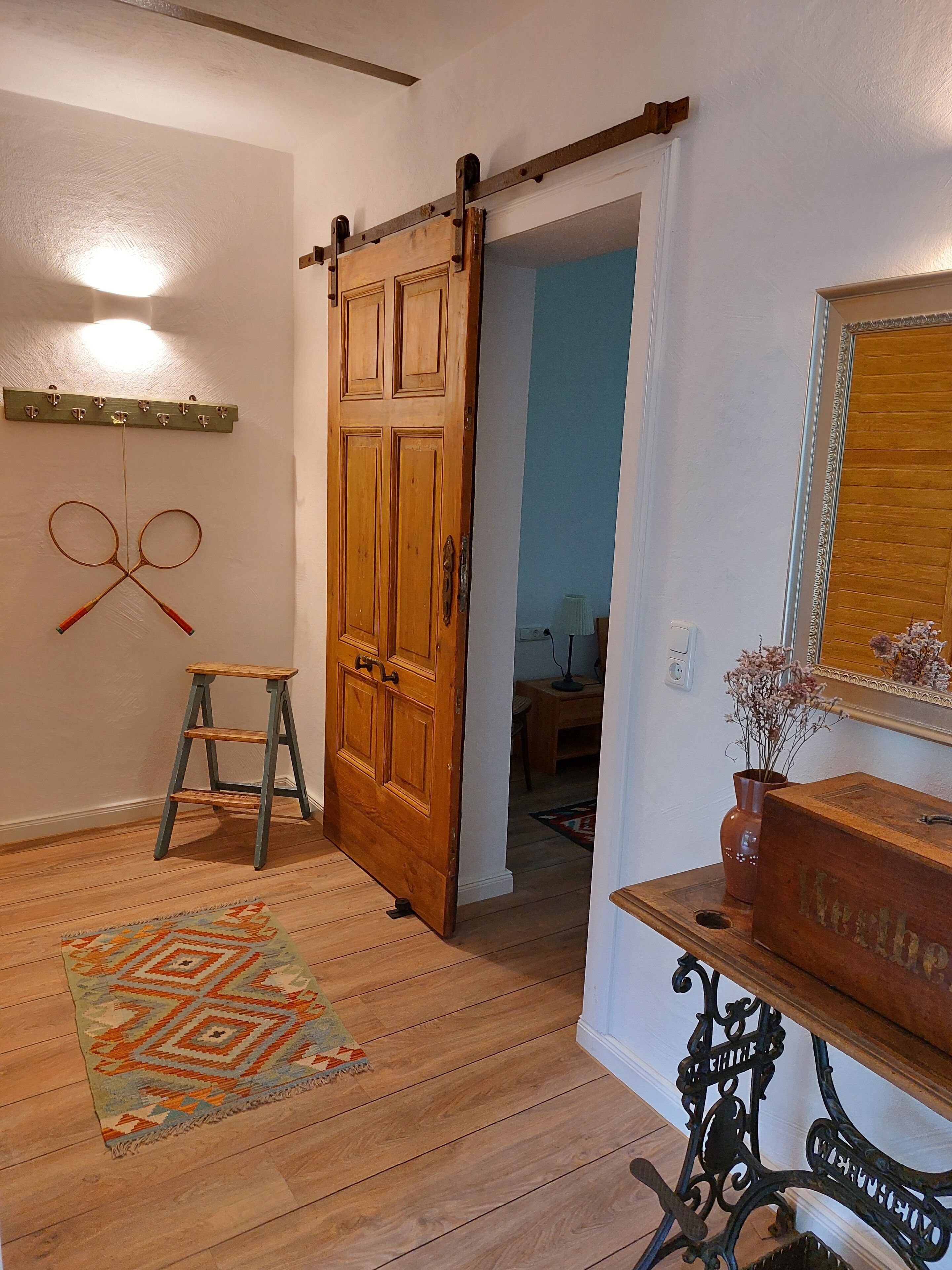 Une pièce avec un plancher en bois, une porte coulissante, un petit tabouret, une table avec un vase et un miroir accroché au mur.