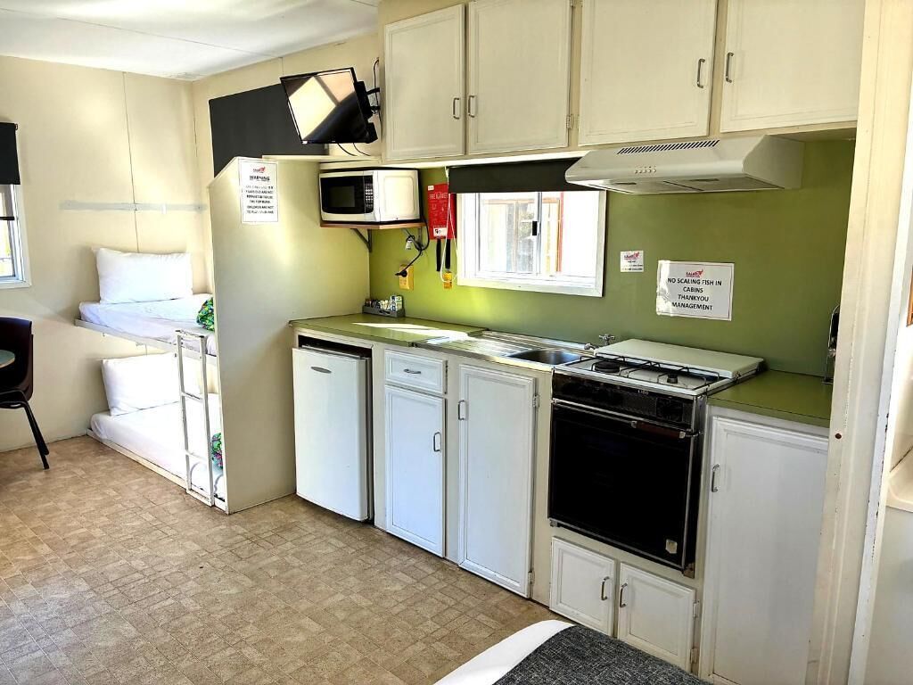 Une cuisine compacte avec des meubles blancs, un four à micro-ondes, une cuisinière et un évier. La pièce adjacente contient un lit avec du linge de lit blanc.