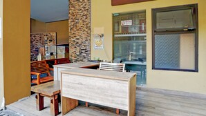 Een houten bank en tafel staan op een houten terras, met een stenen muur en een raam met een bord.