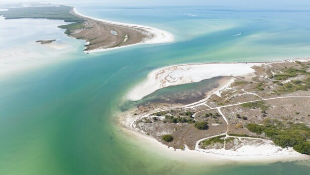 Luchtbeeld van een kustgebied met een zandstrand, helder water en een kleine nederzetting met wegen en begroeiing.