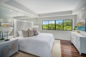 A modern bedroom with a large bed, a nightstand, a dresser, and a large window with a view of greenery. - Hanalei Plantation Villa (Princeville)