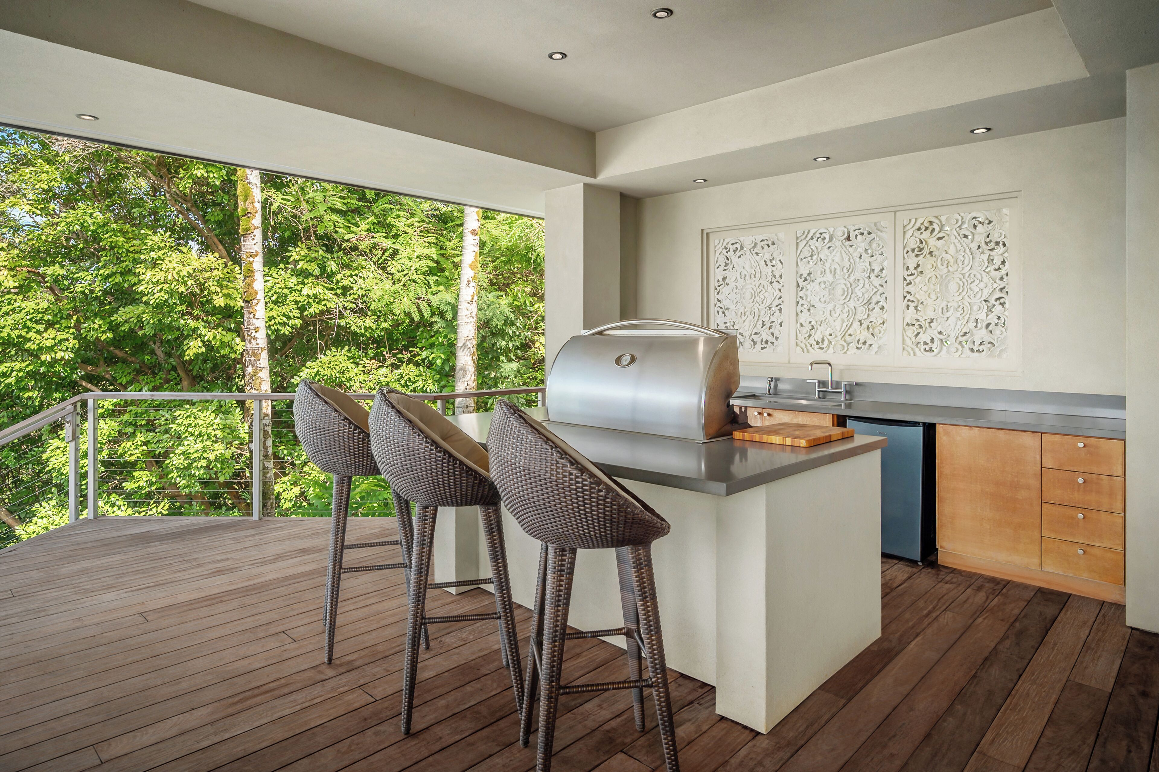 An outdoor kitchen with a grill, two barstools, and a view of trees.