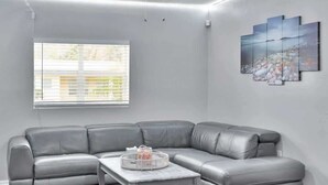 En moderne stue med en grå lædersofa i krogsofa-opstilling, et marmorbord og et stort vindue med persienner.