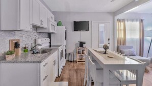 A compact kitchen with white cabinets, a granite countertop, a built-in oven, and a microwave. There's a small dining area with a white table and bench, a globe on the table, and a TV mounted on the wall. - Cozy Suite Full Amenitie s pet Friendly (Tampa)