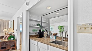 Una cocina moderna con armarios blancos, encimera de granito y pileta de acero inoxidable. Se puede ver un comedor a través de una puerta abierta, con una silla de mimbre y una lámpara.