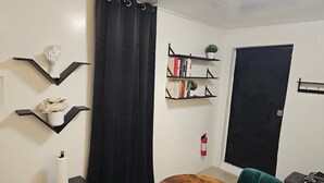 A compact living space with a black curtain, a wooden table, a teal chair, and black shelves with books and a plant. - Ponce Pueblo Central Apt (Ponce)