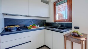 A kitchen with white cabinets, a black countertop, a window with blinds, and a fruit bowl on a small table. - Appartamento Residenza Chiara: Sunny Balcony and Mountain View (Laveno Mombello)