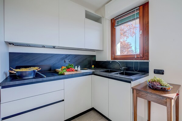 Una cocina con armarios blancos, encimera negra, ventana con persianas y un frutero sobre una mesita.