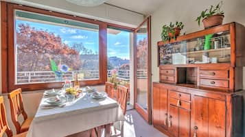 Zona pranzo con tavolo apparecchiato per quattro persone, un mobile contenitore in legno con ante in vetro e vista su alberi autunnali attraverso ampie finestre.