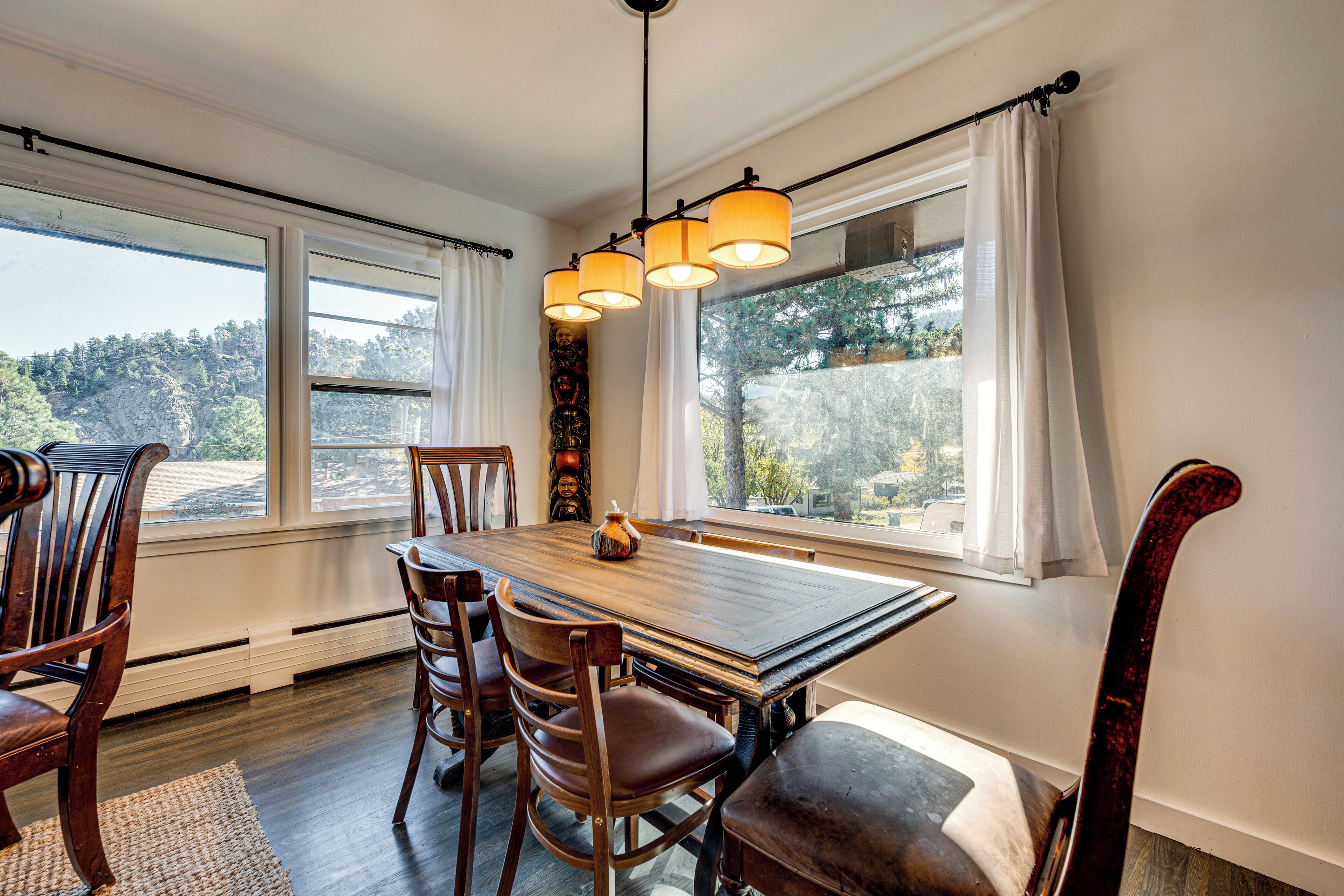 A dining area with a wooden table and chairs, large windows with white curtains, and a hanging light fixture.