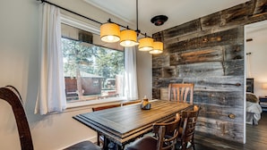 A dining area with a wooden table and chairs, a window with white curtains, and a rustic wooden wall. - Mtn-view Retreat w/ Deck & Hot Tub in Estes Park! (Estes Park)