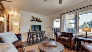 A cozy living room with a leather sofa, a coffee table, and a television. There is a dining area with a table and chairs, and a large window providing a view of the outdoors. - Mtn-view Retreat w/ Deck & Hot Tub in Estes Park! (Estes Park)