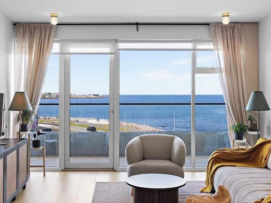 A modern living room with a sofa, a coffee table, and a TV on a cabinet. There is a large sliding glass door leading to a balcony with a view of the sea.
