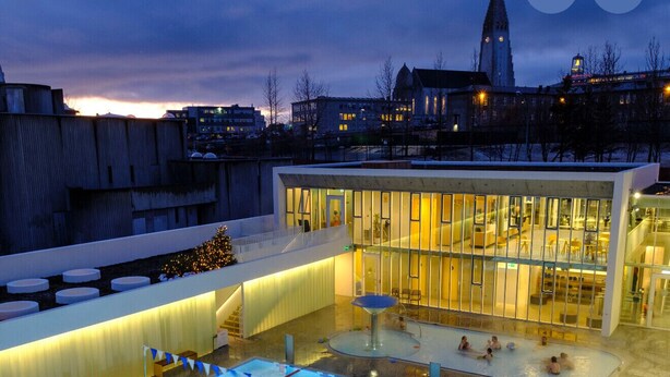 A swimming pool with a glass enclosure, a covered seating area, and a building with large windows. - Ice Apartments (Reykjavík)
