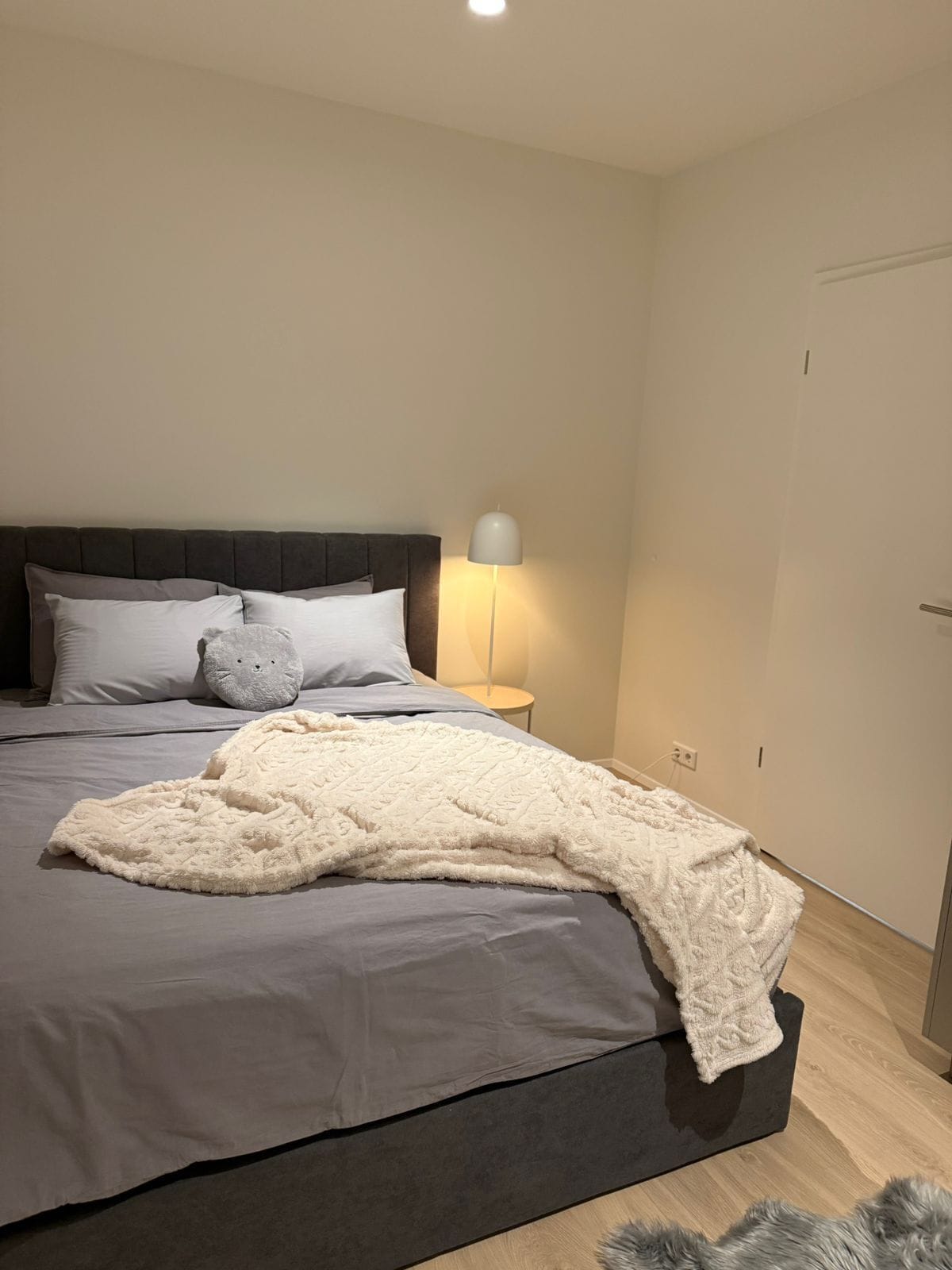 A neatly made bed with a grey bedspread and a white throw blanket, a bedside lamp, and a grey cat-shaped pillow.