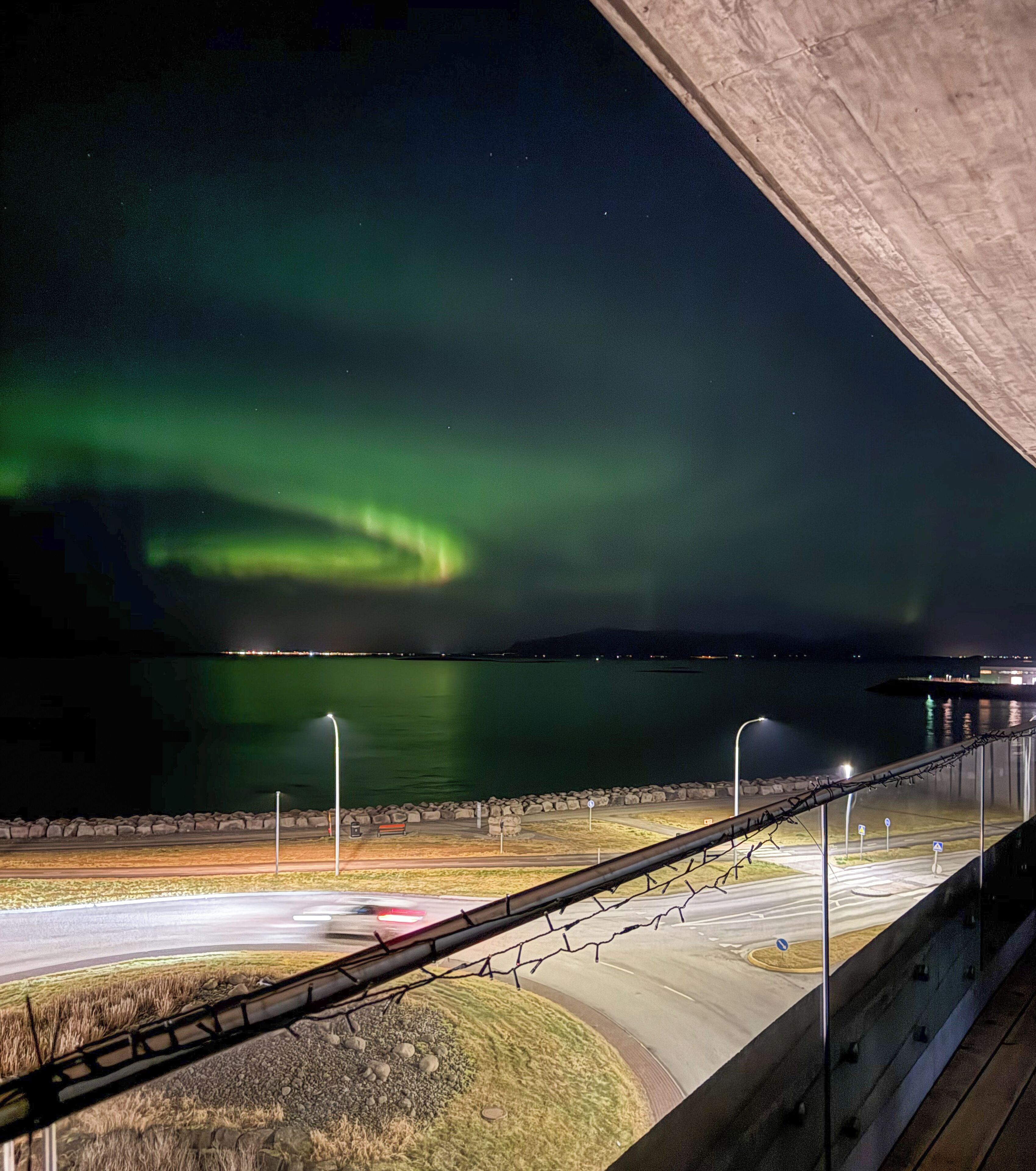 A nighttime view of a bridge with a road beneath, a body of water, and the Northern Lights in the sky.