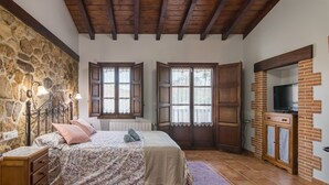 Een slaapkamer met een stenen muur, houten balken in het plafond, een bed met een geborduurd sprei, twee ramen met luiken, een houten kast met een televisie, en een paars vloerkleed.