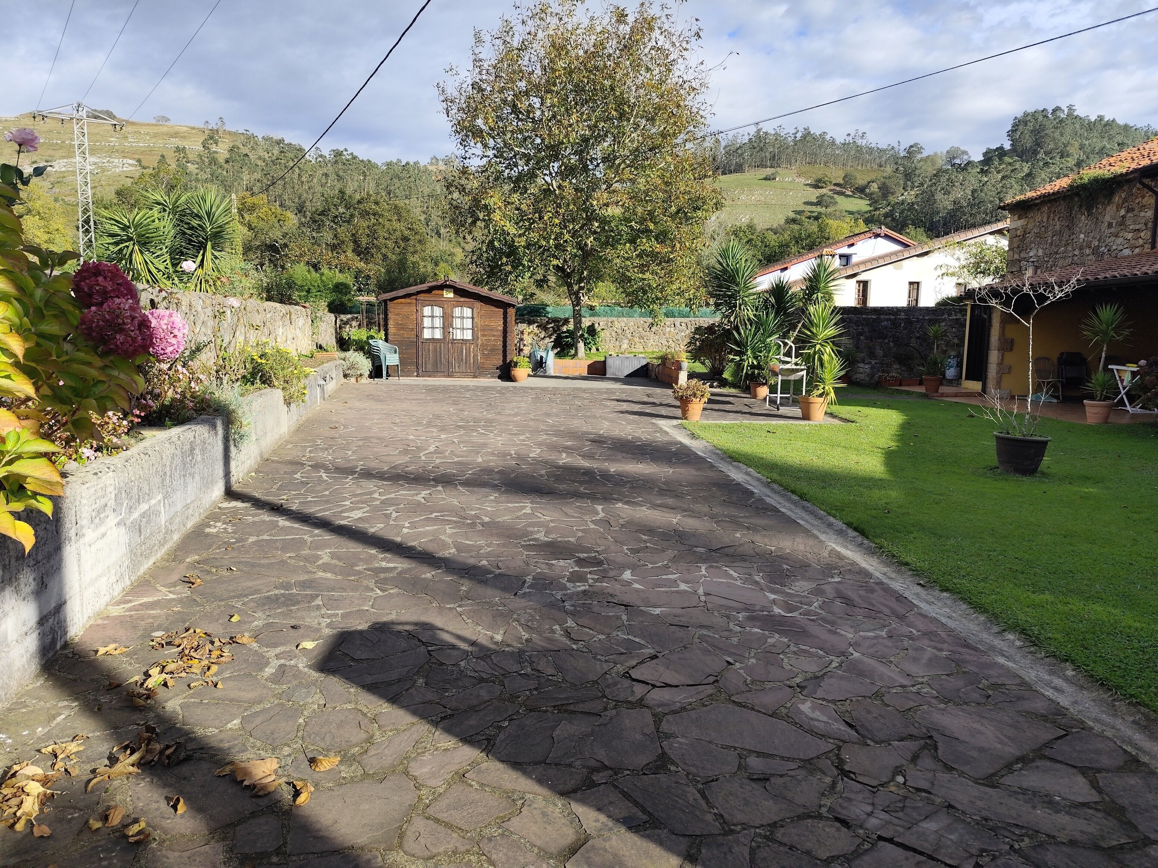 A stone-paved pathway leading to a rustic wooden building with a thatched roof, surrounded by lush greenery and a hilly backdrop.