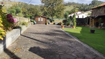 A stone-paved pathway leading to a rustic wooden building with a thatched roof, surrounded by lush greenery and a hilly backdrop.