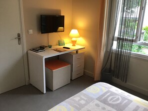 Une chambre à coucher de taille modeste, comprenant un lit simple, un bureau avec une lampe, une télévision et une fenêtre avec des rideaux.