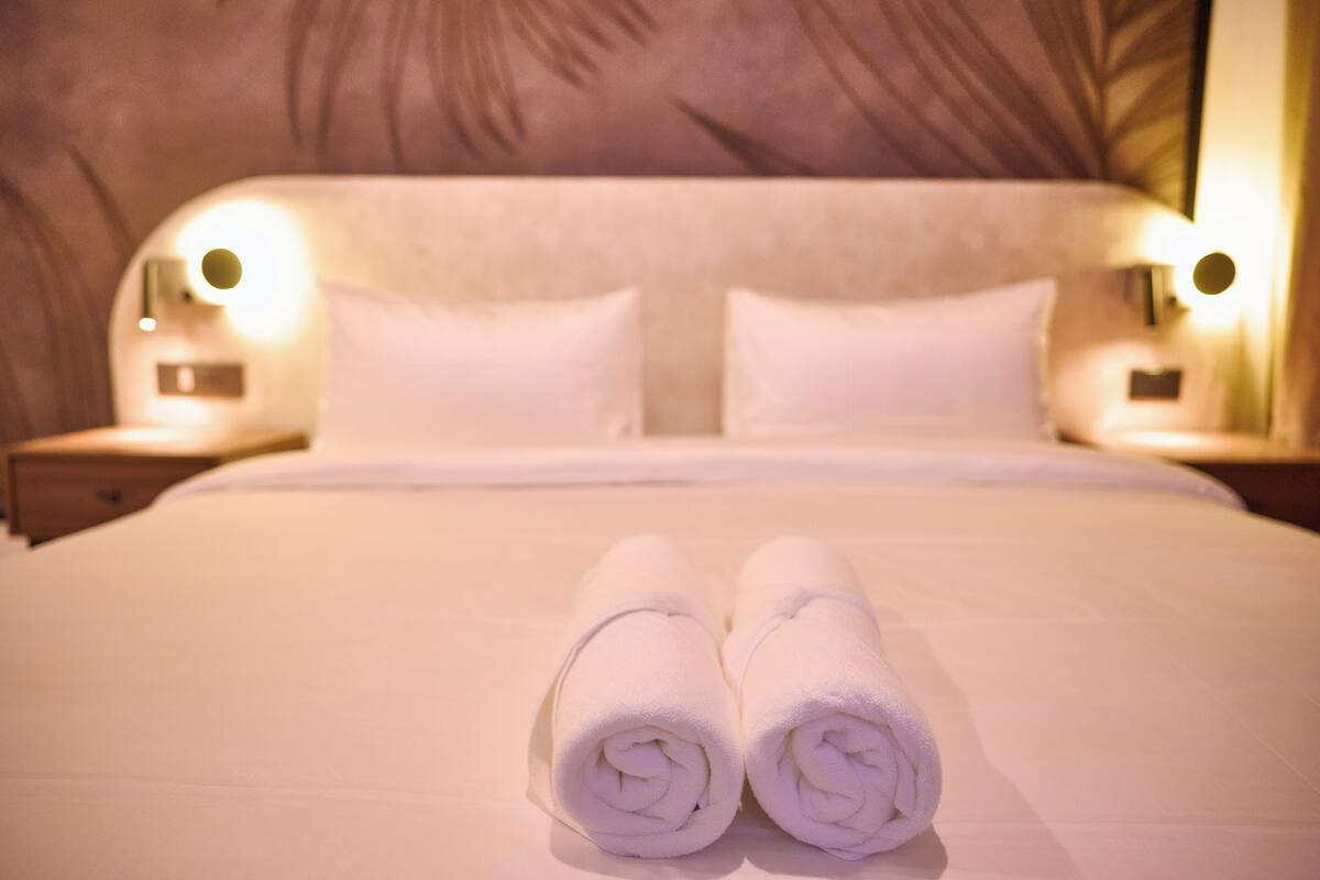 A neatly made bed with two rolled towels in front, a headboard with a textured pattern, and two bedside lamps with round lights.