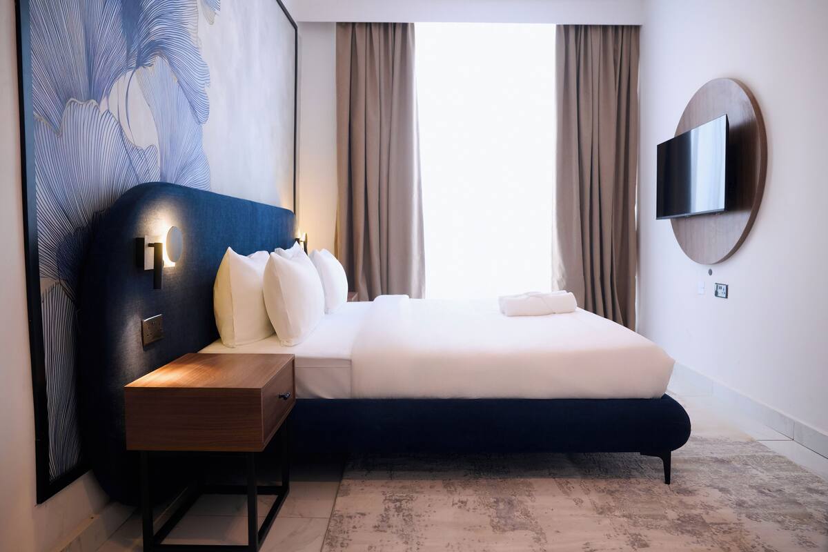 A modern hotel room with a large bed, a bedside table, a wall-mounted TV, and a large abstract painting.