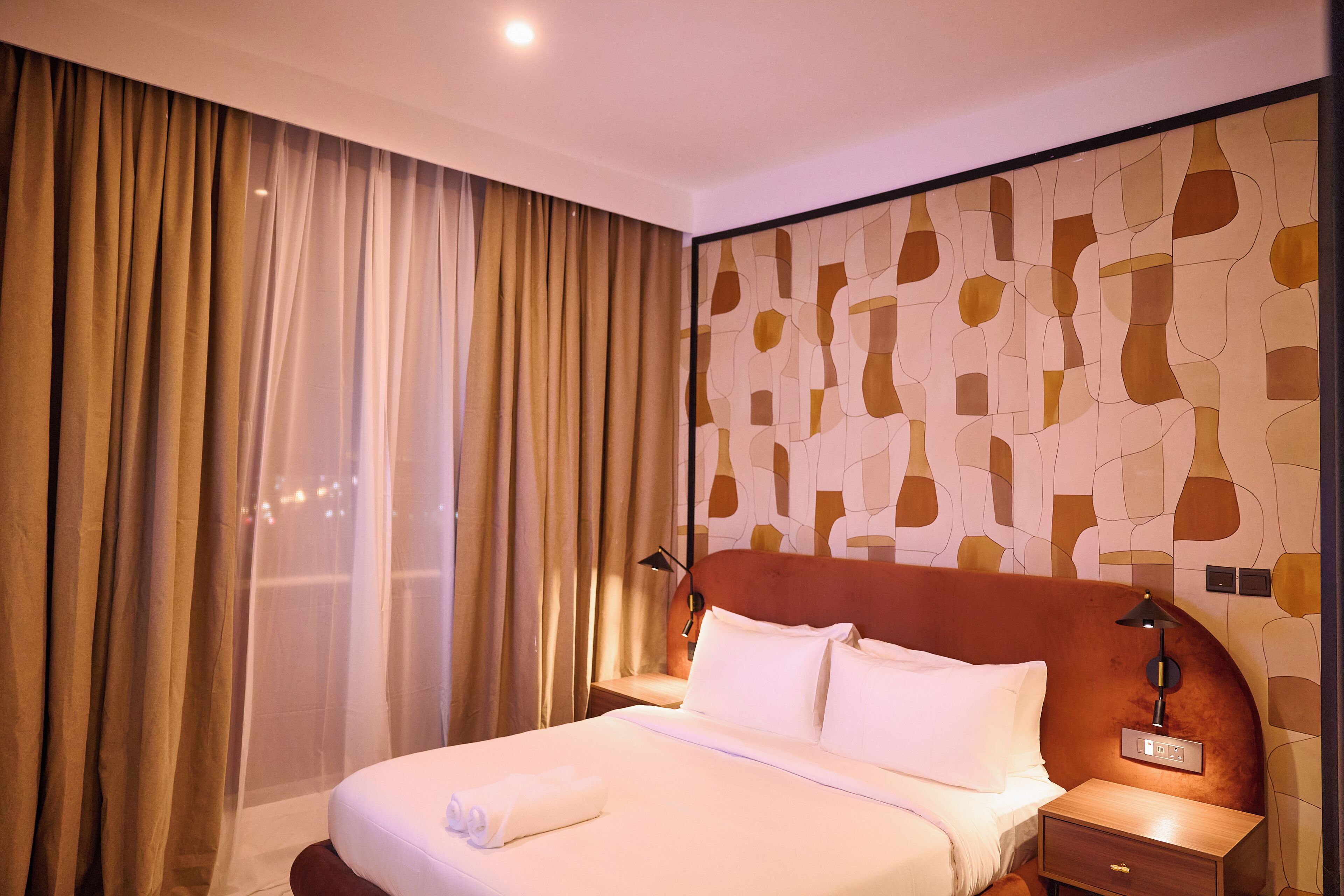 A hotel room with a large bed, a nightstand, a lamp, and a view through sheer curtains.